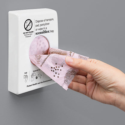 Hand using a wall-mounted dispenser to retrieve a scensibles bag for menstrual products on a gray background.